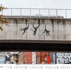 Text: "Oktober", "Three Monkeys, 2024, Brick Lane, London". Eine Brücke mit gemalten Affen und darunter ein urbanes Straßenszenario.