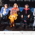 Text: "OKTOBER", Kalenderblatt mit Wochentagen und Daten. Darunter sitzen vier Personen auf Stühlen, fröhlich und elegant gekleidet.