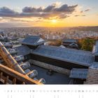 Blick von der Burg Matsuyama bei Sonnenuntergang. Im Vordergrund traditionelle Gebäude, im Hintergrund die Stadt und Berge.