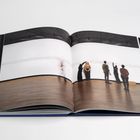 Keine sichtbaren Texte. Geöffnetes Buch zeigt Personen, die in einer Galerie eine minimalistische Wandinstallation betrachten.