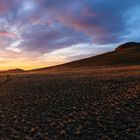 Eine weite Landschaft bei Sonnenuntergang, hügeliges Gelände mit Gras, leuchtender Himmel in Orange und Blau.