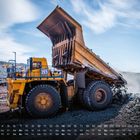 Kalender mit Datum und Tagen unten, großes gelbes Muldenkipperfahrzeug kippt Schutt auf einem unbebauten Gelände ab.