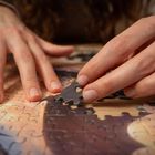 Zwei Hände legen ein Puzzle, eines der Teile wird gerade platziert. Man sieht die Details der Haut und Nägel.