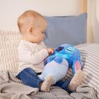Ein Kleinkind in einem weißen Shirt und blauen Jeans hält ein blaues Plüschtier auf einem Bett mit Kissen.