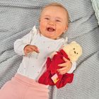 Ein lächelndes Baby liegt auf einer grauen Decke und hält ein Kuscheltier mit roter Kleidung.