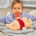 Ein Baby mit blauem Hemd beißt in einen roten Beißring und liegt auf einer grauen Decke neben einem Plüschbären.