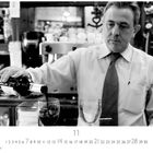 Ein Mann in Hemd und Krawatte schenkt Rotwein in ein Glas ein. Im Hintergrund sind Flaschen und eine Espressomaschine zu sehen.