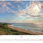 "Wolkenlandschaft am Wüstenstrand" zeigt einen Strand mit grünem Ufer, teils bewölkten Himmel und Sonnenstrahlen über dem Meer.