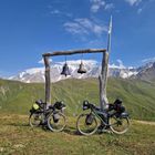 Eine Glocke mit der Aufschrift "1993" hängt an einem Holzgestell. Zwei bepackte Fahrräder darunter, im Hintergrund Berge.