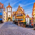 Kopfsteinpflasterstraße in einer Altstadt. Fachwerkhäuser in Gelb und Blau. Ein Tor mit Turm und Uhr im Hintergrund.
