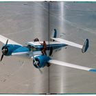 Text: "N75WB". Ein Mann klettert auf einem fliegenden, zweimotorigen Propellerflugzeug. Wüstenlandschaft im Hintergrund.