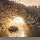11 | November. Ein natürlicher Steinbogen über einem ruhigen Fluss, beleuchtet von warmem Sonnenlicht.