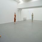 Vier schlanke Figuren in farbigen Kleidern aus hellem Material stehen in einem leeren, weißen Raum.