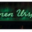 „Blumen Ursprung“ steht in grüner Neonschrift auf dunklem Hintergrund, eine Werbeaufschrift.