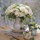 Ein Strauß weißer Rosen in einer weißen Vase auf einem Tisch mit Häkeldeckchen, umgeben von kleineren Blumenarrangements.