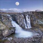 Der Vollmond leuchtet über einem Wasserfall, der über eine felsige Klippe in einen ruhigen Pool stürzt.