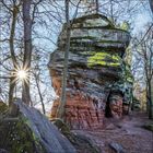 Ein großer Sandsteinfelsen in einem Wald, umgeben von kahlen Bäumen. Die Sonne scheint grell durch die Äste.