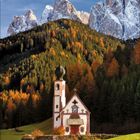 November 2023. Eine kleine Kirche vor einem herbstlichen Wald, dahinter schneebedeckte Berge und blauer Himmel.