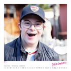 Novemberkalender, Juliana, lächelnde Person mit roter Mütze, hält ein kleines Gerät, Hintergrund grün.