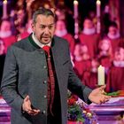 Ein Mann in Tracht gestikuliert vor einem Altar, im Hintergrund ein Chor in roten Roben und brennende Kerzen.