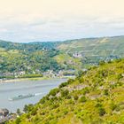 Text "DER RHEIN" oben und "Am Mittelrhein" unten. Ein Fluss mit grün bewachsenen Hügeln und einem fahrenden Schiff.