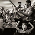 Menschen jubeln und reißen die Hände hoch in einer vollen Bar. Ein Kind in gestreiftem Shirt schreit begeistert.