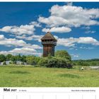 Mai 2027, Kalender, Woche 21. Foto eines hölzernen Turms auf einer grünen Wiese, umgeben von Bäumen unter blauem Himmel.