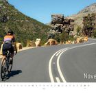 Text: "November". Ein Radfahrer auf einer kurvenreichen Bergstraße, umgeben von grünen Hügeln und steinigen Felsen.