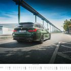 "Oktober" steht unten. Ein grüner BMW-Kombi fährt auf einer Straße, daneben eine Leitplanke, blauer Himmel.