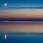 Oben ein heller, schmaler Mond. Darunter ein stiller See mit pastellfarbenem Himmel im Hintergrund.