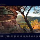 09 September. Ein großer Felsen und ein Baum im Vordergrund mit Blick auf ein sonniges Waldtal im Hintergrund.