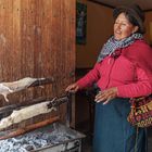Eine Frau in traditioneller Kleidung steht neben einem Grill mit zwei gebratenen Tieren. Holzverkleidung im Hintergrund.