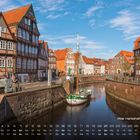 „Alter Hansehafen, Stade 11“ in weißer Schrift unten rechts. Fachwerkhäuser, Kanal und Segelboot unter blauem Himmel.
