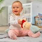 Keine Texte vorhanden. Ein Baby in einem hellen Raum hält ein Plüschtier und sitzt auf Decken.