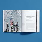Text: "Raising the Flag at Ground Zero." Drei Feuerwehrleute hissen die US-Flagge. Ruinen sind im Hintergrund sichtbar.