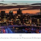"Skyline 12" und "CASARES EDITION". Abendstimmung mit beleuchteter Stadt, Hafen und Schiffen; Sonnenuntergang am Horizont.