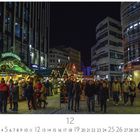 Weihnachtsmarkt, Entenbraterei. Menschen flanieren zwischen hell erleuchteten Ständen und modernen Gebäuden bei Nacht.