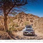 Ein Auto mit "CURVYTAN"-Schriftzug auf einer staubigen, gebirgigen Straße, links ein Baum, rechts ein Lehmhaus.