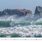 Unten steht "12" und eine Monatsübersicht. Ein Windsurfer springt über eine große, stürmische Welle vor Felsen im Hintergrund.