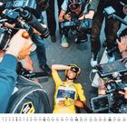 Kalender unten mit Zahlen von 1 bis 31. Radfahrer in gelbem Trikot liegt, umgeben von Fotografen mit Kameras.