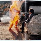 Eine Person in Flammen steht nahe einem Mann in Anzug, der auf einem Felsen sitzt, umgeben von karger Landschaft.