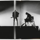 Zwei Silhouetten: Links steht jemand mit Gitarre, rechts sitzt eine Person auf einem Stuhl, in starkem Bühnenlicht.