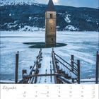 Kalender für Dezember mit verschneitem Steg und halb untergegangenem Kirchturm vor einer Bergkulisse.