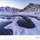Text: "DEZEMBER". Schneebedeckte Berglandschaft mit gefrorenem See, im Hintergrund eine beleuchtete Siedlung.