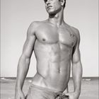 "12" und ein Kalendermonat darunter. Ein muskulöser Mann in Shorts steht am Strand. Schwarz-Weiß-Fotografie.