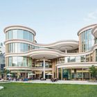 Moderne, geschwungene Glasfassade mit runden Formen, umgeben von grüner Landschaft und Menschen im Außenbereich.