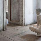 Keine Texte sichtbar. Eine Skulptur eines sitzenden Menschen in einem Raum mit Holzboden und abgenutzten Wänden.