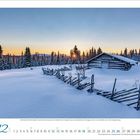 12 Dezember. Winterlandschaft mit Holzhaus, verschneiter Zaun vor Sonnenuntergang. Bäume im Hintergrund unter klarem Himmel.