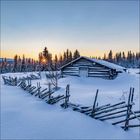 Holzhütte und Zaun im Schnee, Tannen im Hintergrund, Sonnenuntergang am klaren Himmel.