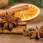 Anisstern, Zimtstangen und eine Orangenscheibe liegen auf einem Holztisch. Im Hintergrund sind Tannenzweige erkennbar.